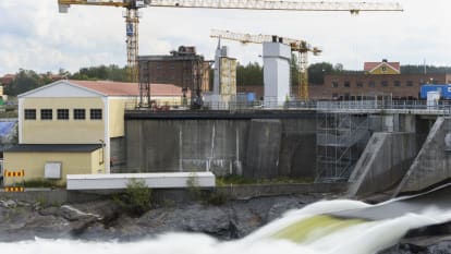 Bild på en vattenkraftstation, där vatten forsar i en flod samtidigt som lyftkranar, bostadshus och skog syns i bakgrunden.