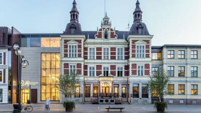 Picture of the Kristianstad city hall, with red and white facade, castle-like rooftops, and a modern concrete entrance adjoined to the left.