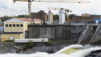 Bild på en vattenkraftstation, där vatten forsar i en flod samtidigt som lyftkranar, bostadshus och skog syns i bakgrunden.