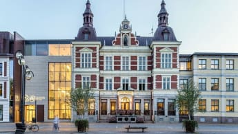 Picture of the Kristianstad city hall, with red and white facade, castle-like rooftops, and a modern concrete entrance adjoined to the left.