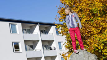 En flicka står på en sten, med ett vitt radhus åt vänster bakgrund och ett höstfärgat träd åt höger bakgrund.