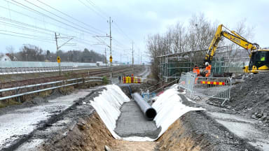 Rörläggning längs med E45. Mitt i bilden syns ett rörschakt med markduk, makadam och ett rör. Till höger om rörschakten står en gul grävmaskin. Till vänster om rörschaktet skymtar man E45.