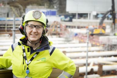 Porträtt på kvinnlig medarbetare iklädd varselkläder och hjälm. I bakgrunden syns arbetsplats med gula stämp.