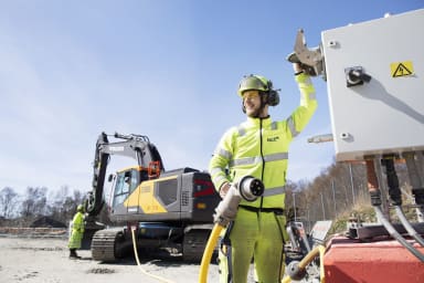 Byggarbetsplats. I förgrunden står en byggnadsarbetare i varselkläder och kopplar in en stor kontakt i ett flyttbart kabelskåp. Kontakten sitter i en kabel som går till en grävmaskin i bakgrunden.