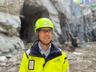 Medarbetare iklädd varselkläder, skyddsglasögon och hjälm blickar utöver arbetsplatsen. I bakgrunden syns tunnelöppningar. Man ser att det är jobb som pågår.