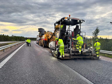Bilden visar en väg som sträcker sig framåt och kröker svagt åt höger. På högra sidan av vägen i framkant av bilden ser man en asfaltläggare där ett antal medarbetare arbetar iklädda varselkläder.