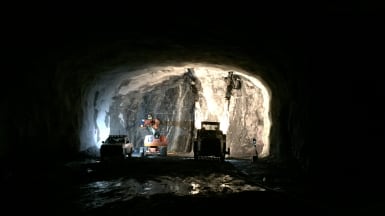 I en delvis upplyst bergtunnel arbetar byggmaskiner, med andra fordon närvarande.