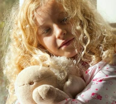 A child leans against a window while holding a bear.