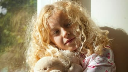 En ung flicka håller i sin nalle, medan hon lutar sig mot ett fönster där solen skiner igenom.