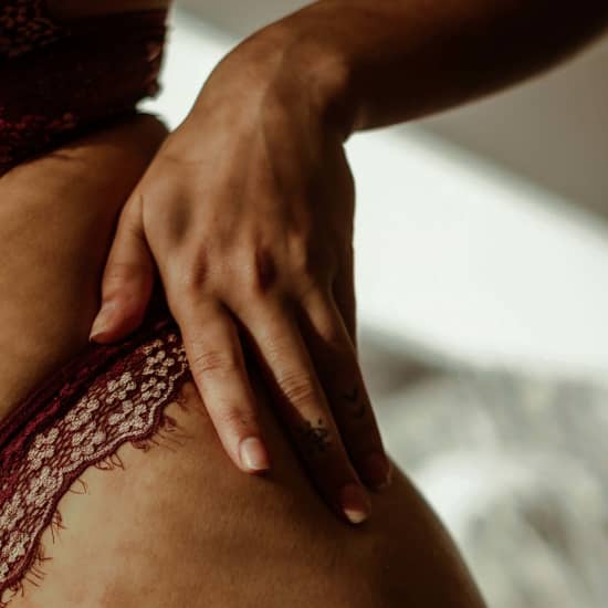 A close-up of a woman’s hand resting gently on her hip, showcasing smooth skin and natural curves. She is wearing burgundy lace lingerie, and a small, minimalistic tattoo is visible on one of her fingers. The soft, natural lighting emphasizes skin texture and highlights the body's contours, complementing a discussion on body sculpting and liposuction outcomes.