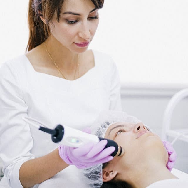 A woman lies calmly on a treatment bed in a modern medspa as a licensed aesthetician performs a laser facial treatment. The aesthetician holds a sleek laser device, gently guiding it across the woman's cheek. Soft lighting highlights the woman's relaxed expression and the clean, clinical setting. The atmosphere is serene and professional, emphasizing advanced skincare and self-care.