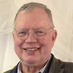 A smiling older man wearing glasses and a striped shirt, with a gray jacket, appears against a light-colored background.