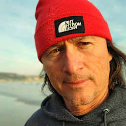 A man with long hair and a beard wearing a red beanie with a logo, standing in front of a body of water with boats and buildings in the background.