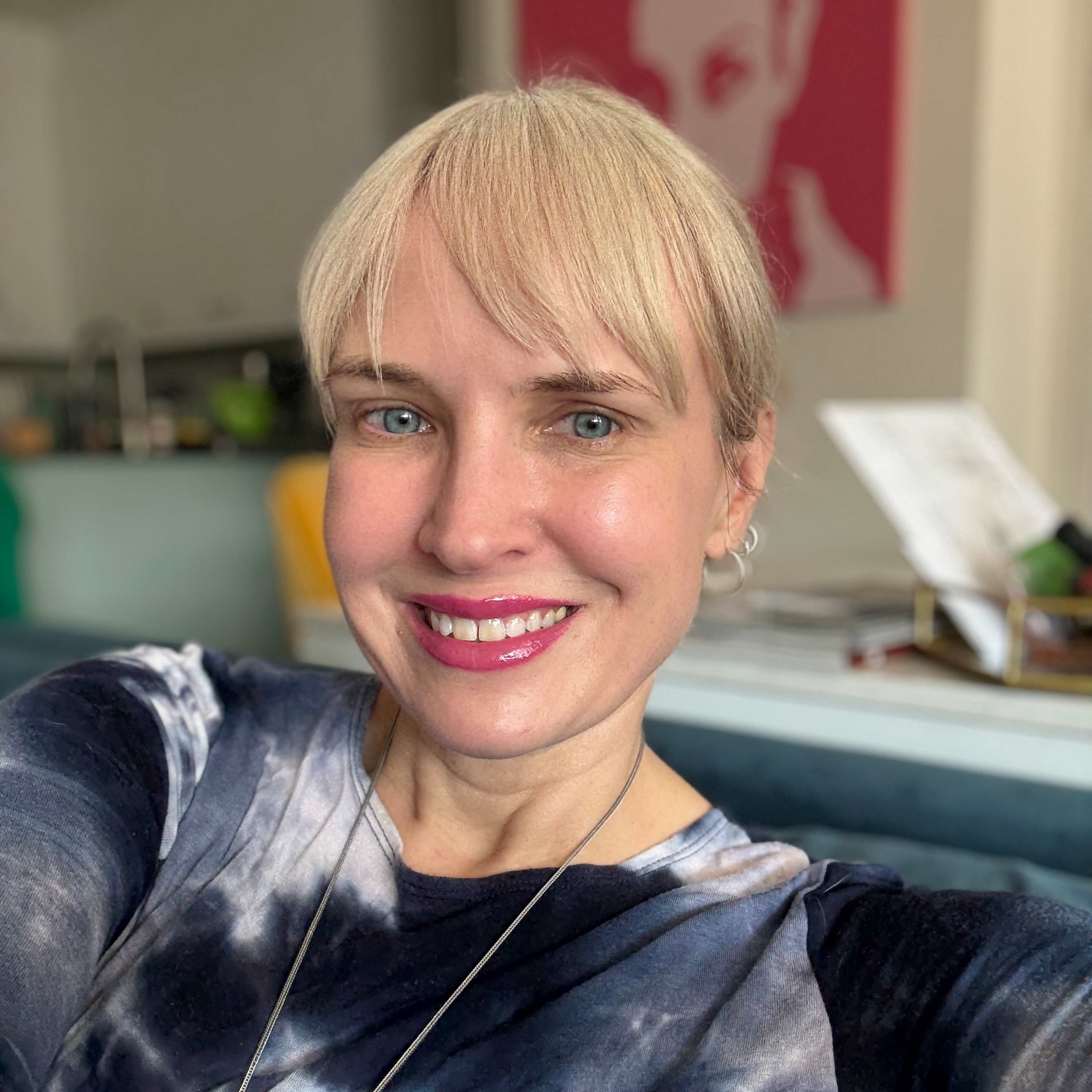 A smiling woman with short blonde hair sits in a room with colorful artwork on the walls.