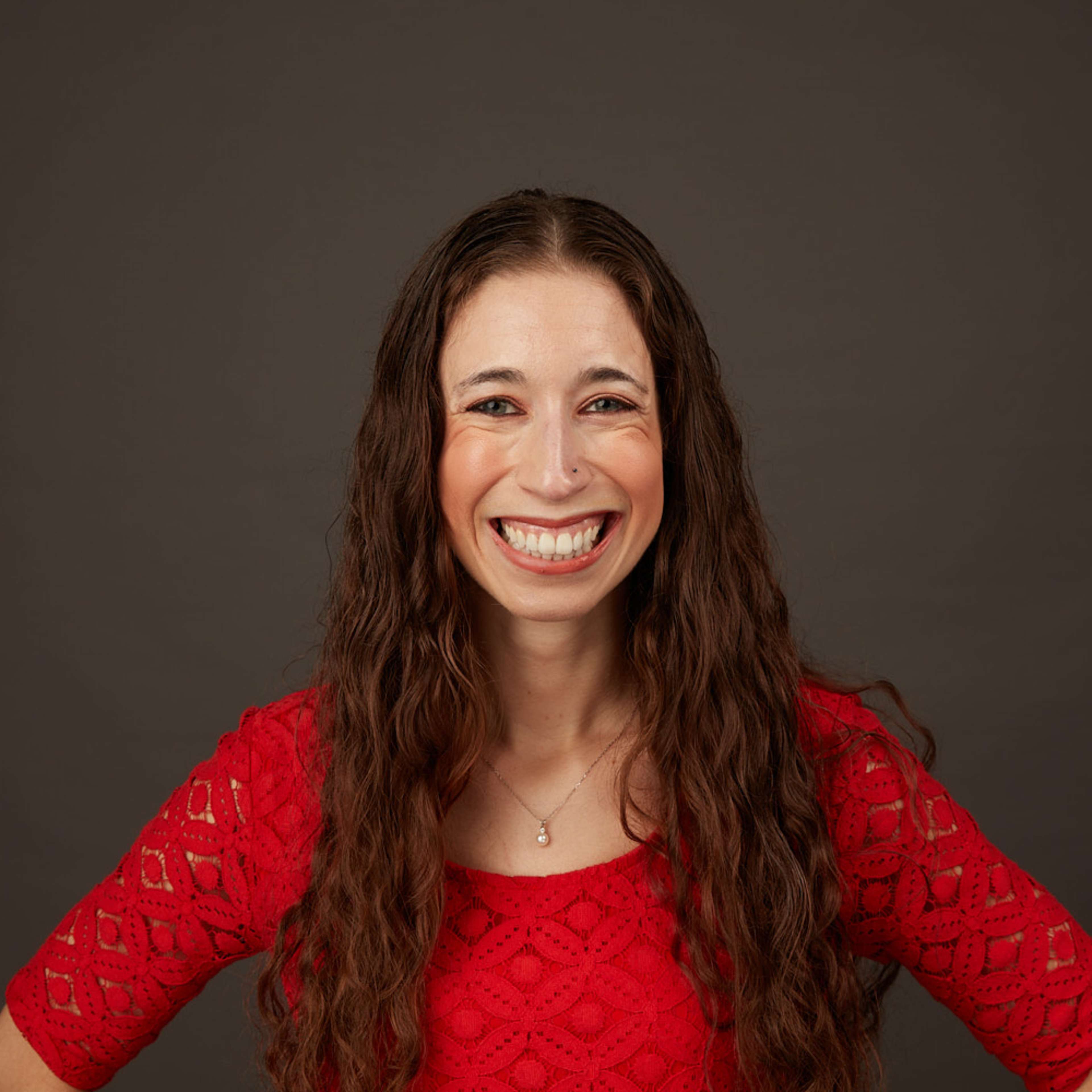 A smiling young woman with long brown hair wearing a red lace top stands against a dark background.