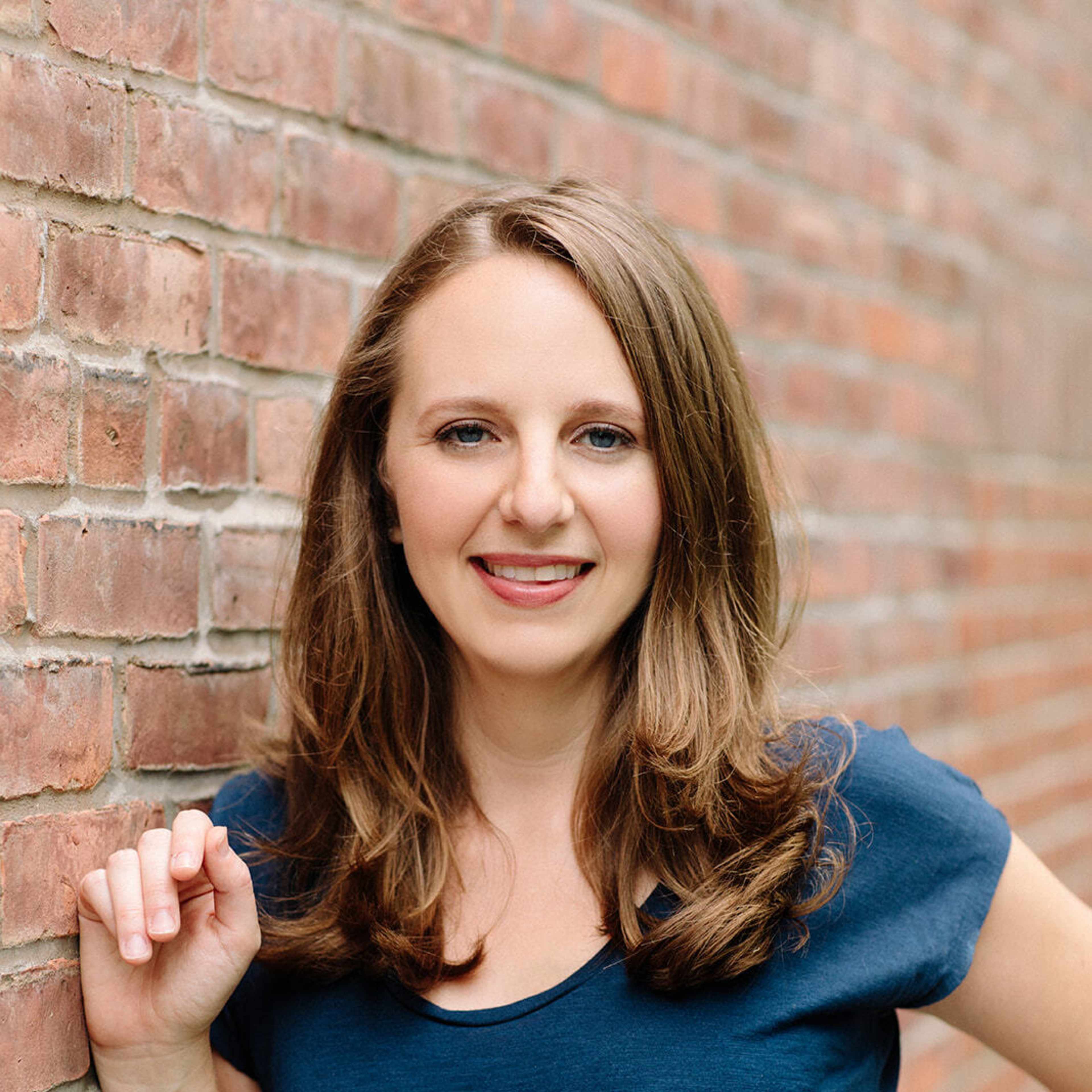 A smiling young woman with curly brown hair stands in front of a brick wall, wearing a blue top.
