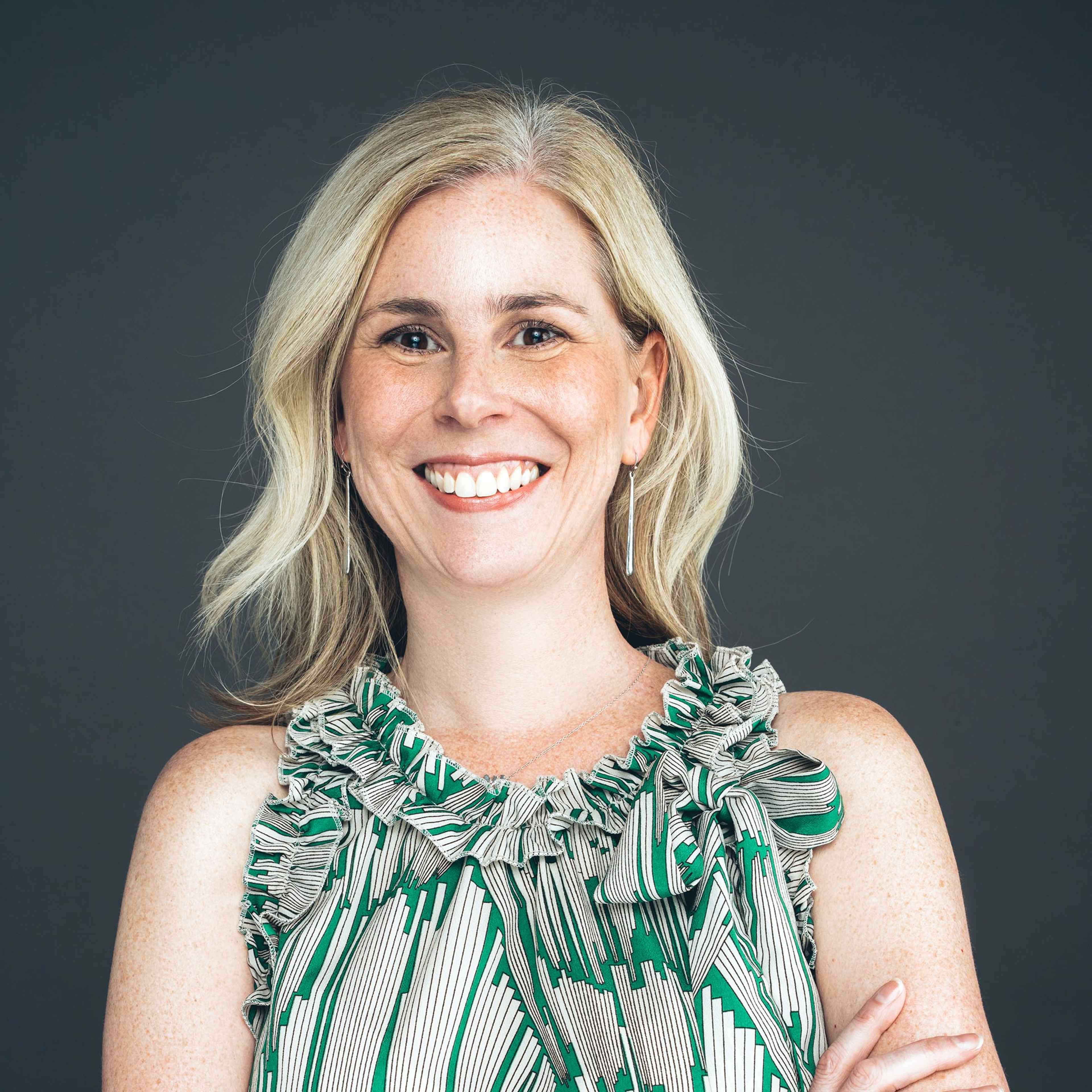 A smiling woman with blonde hair wearing a green and white patterned dress stands against a dark background.
