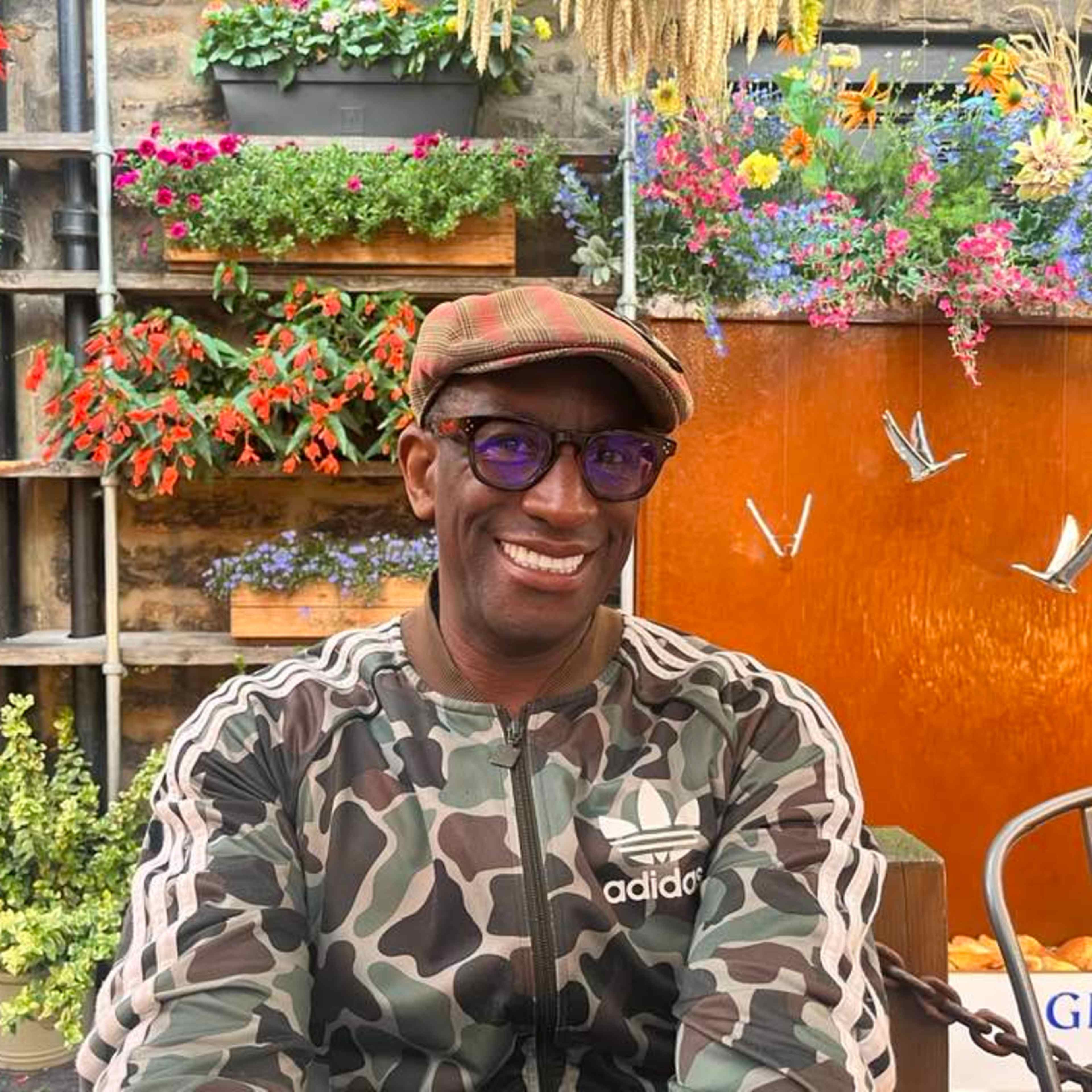 A smiling man wearing a camouflage-patterned shirt and a newsboy cap sits in front of a vibrant, colorful garden display with various potted plants and flowers.