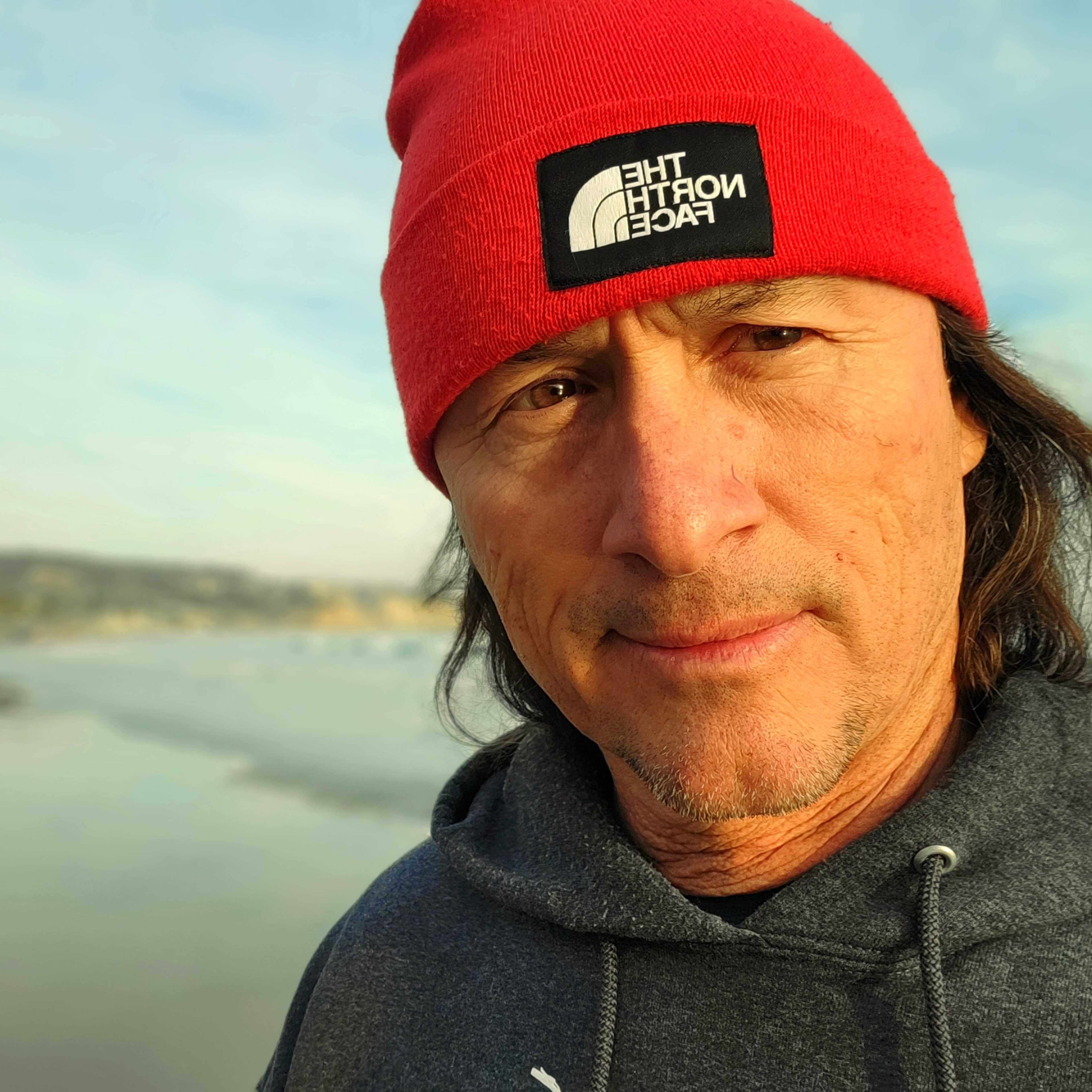 A man with long hair and a beard wearing a red beanie with a logo, standing in front of a body of water with boats and buildings in the background.