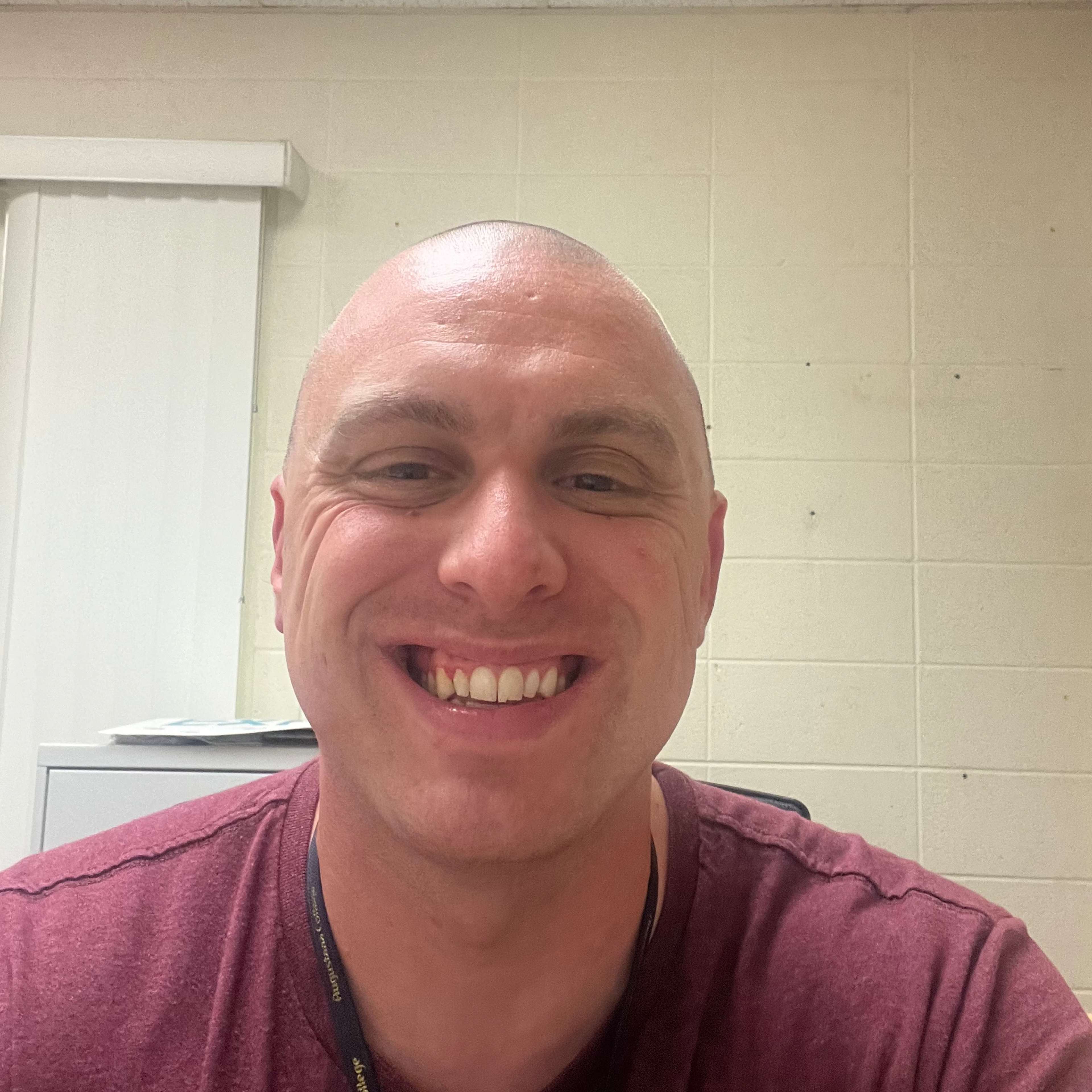 The image shows a smiling bald man wearing a burgundy shirt against a plain background.