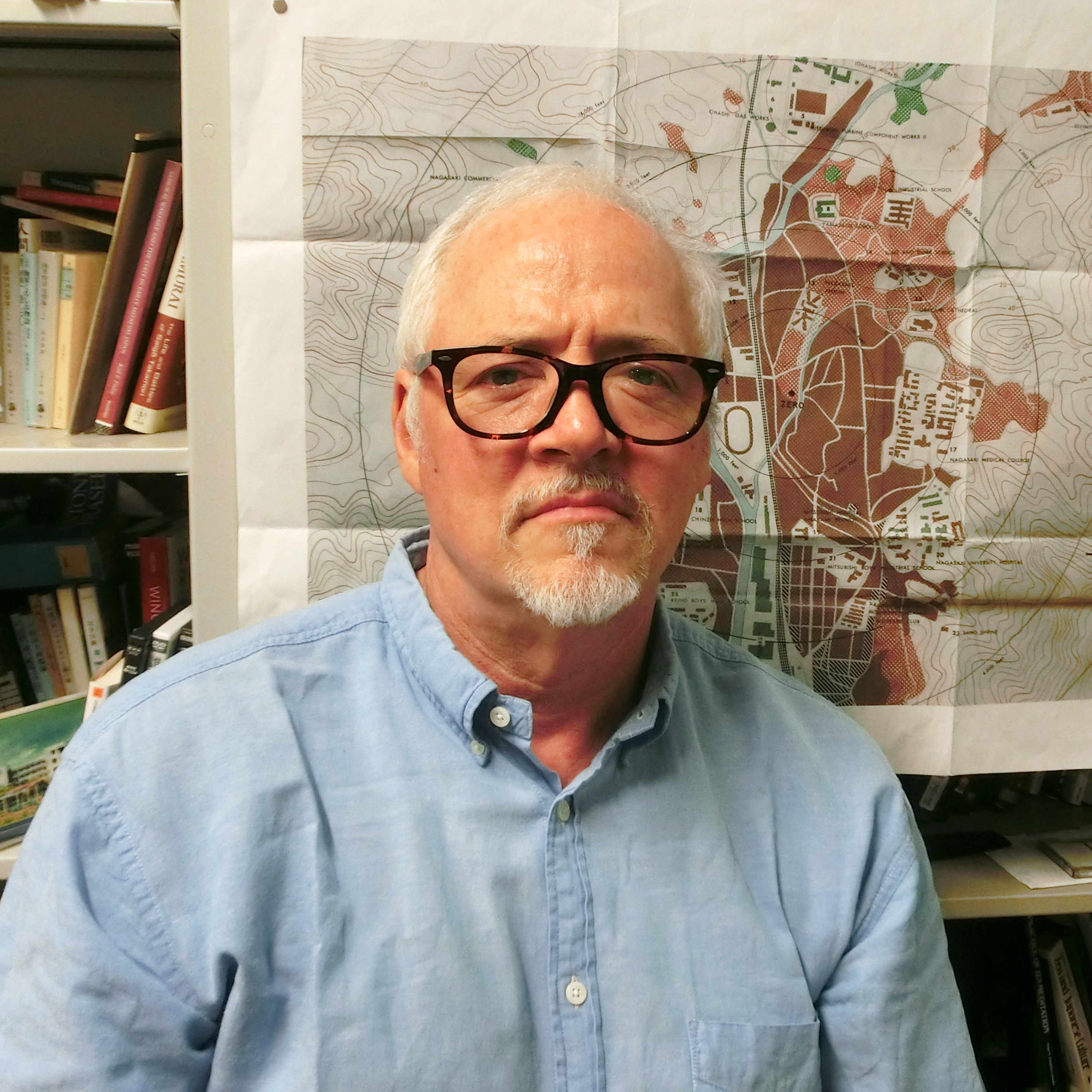 M.G. Sheftall: An older man with gray hair and glasses sits in front of a bookshelf, with a map visible in the background.