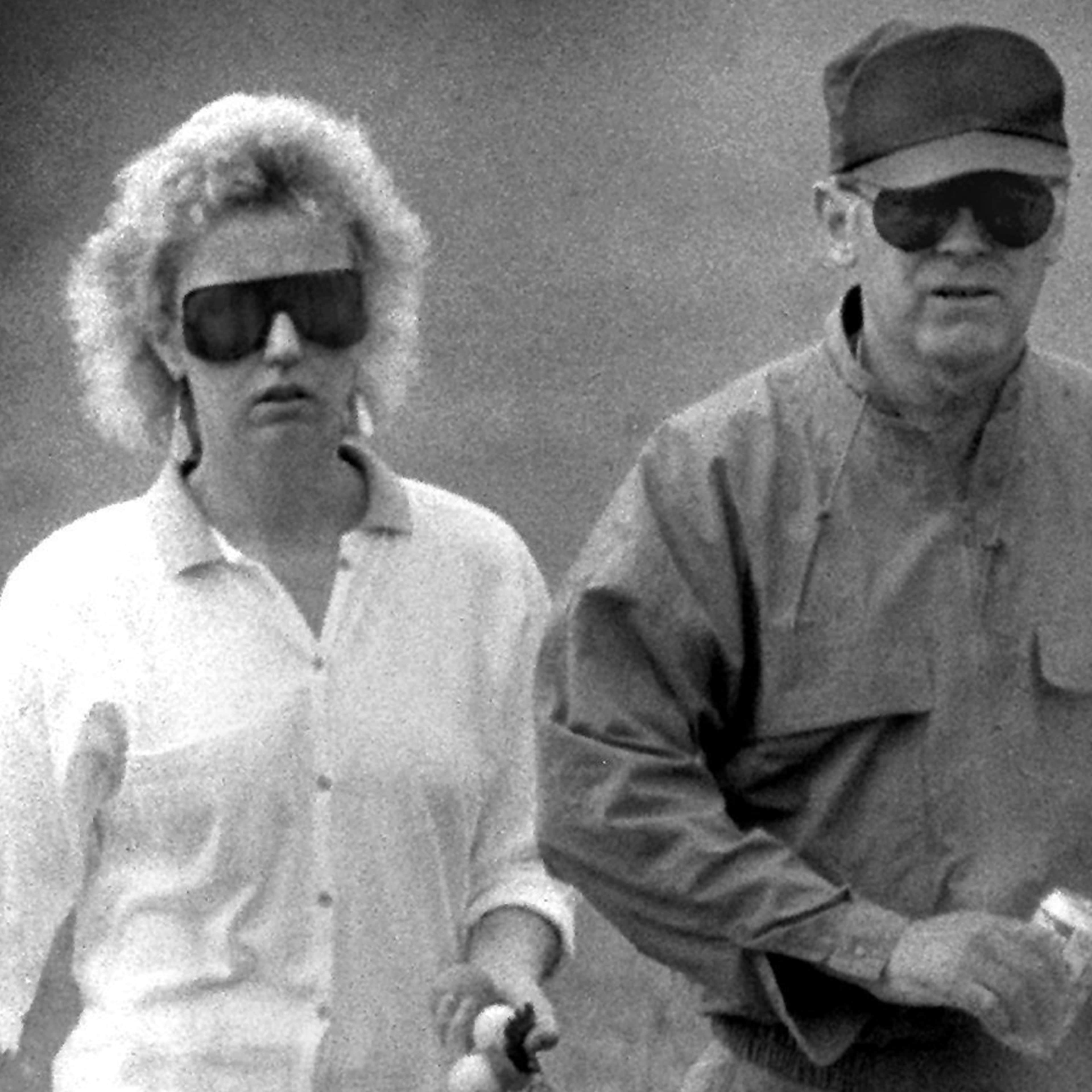 Two individuals, a man and a woman, wearing sunglasses and casual attire, standing together in a black and white photograph.