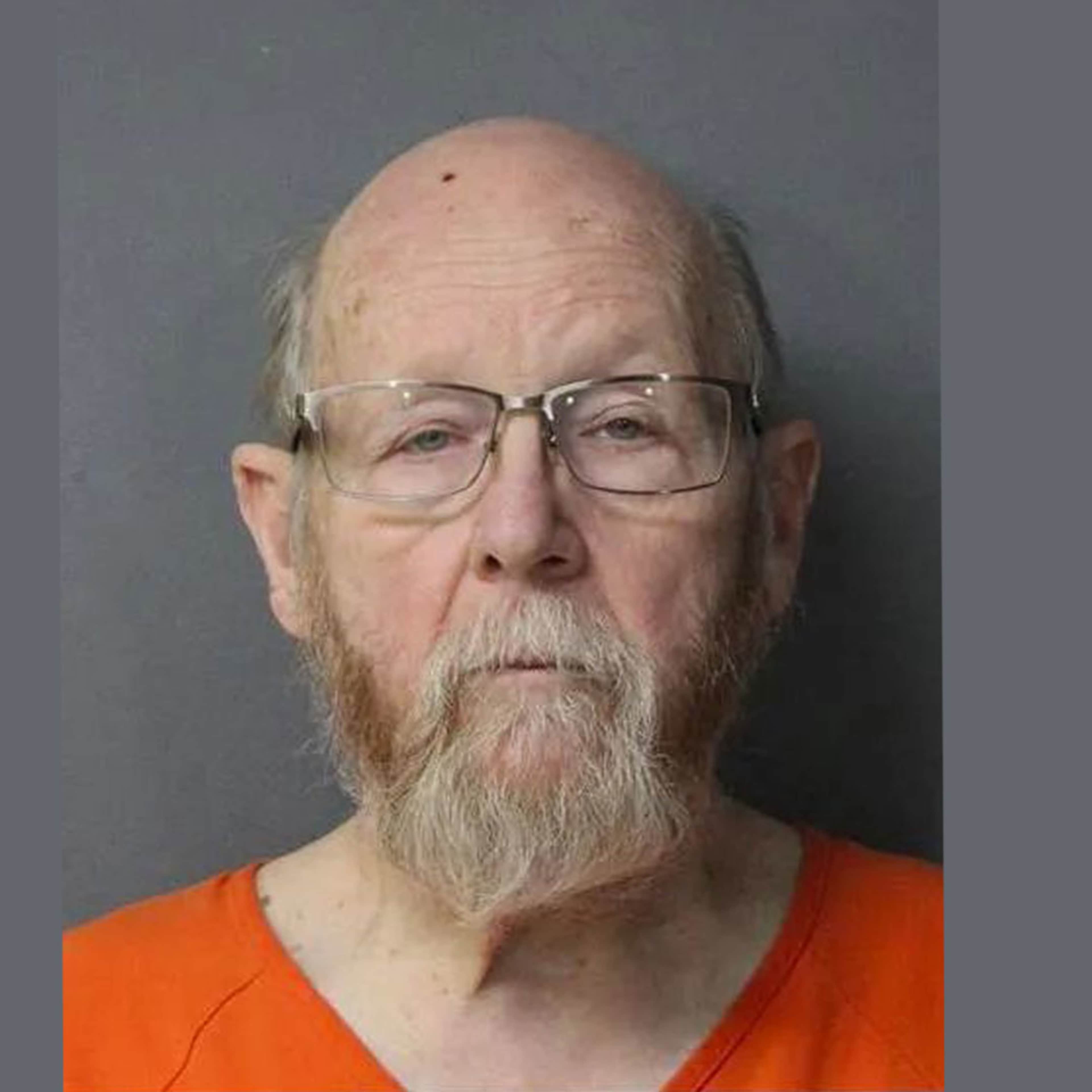 The image shows an elderly man with a long white beard and glasses, wearing an orange prison uniform, against a plain gray background.
