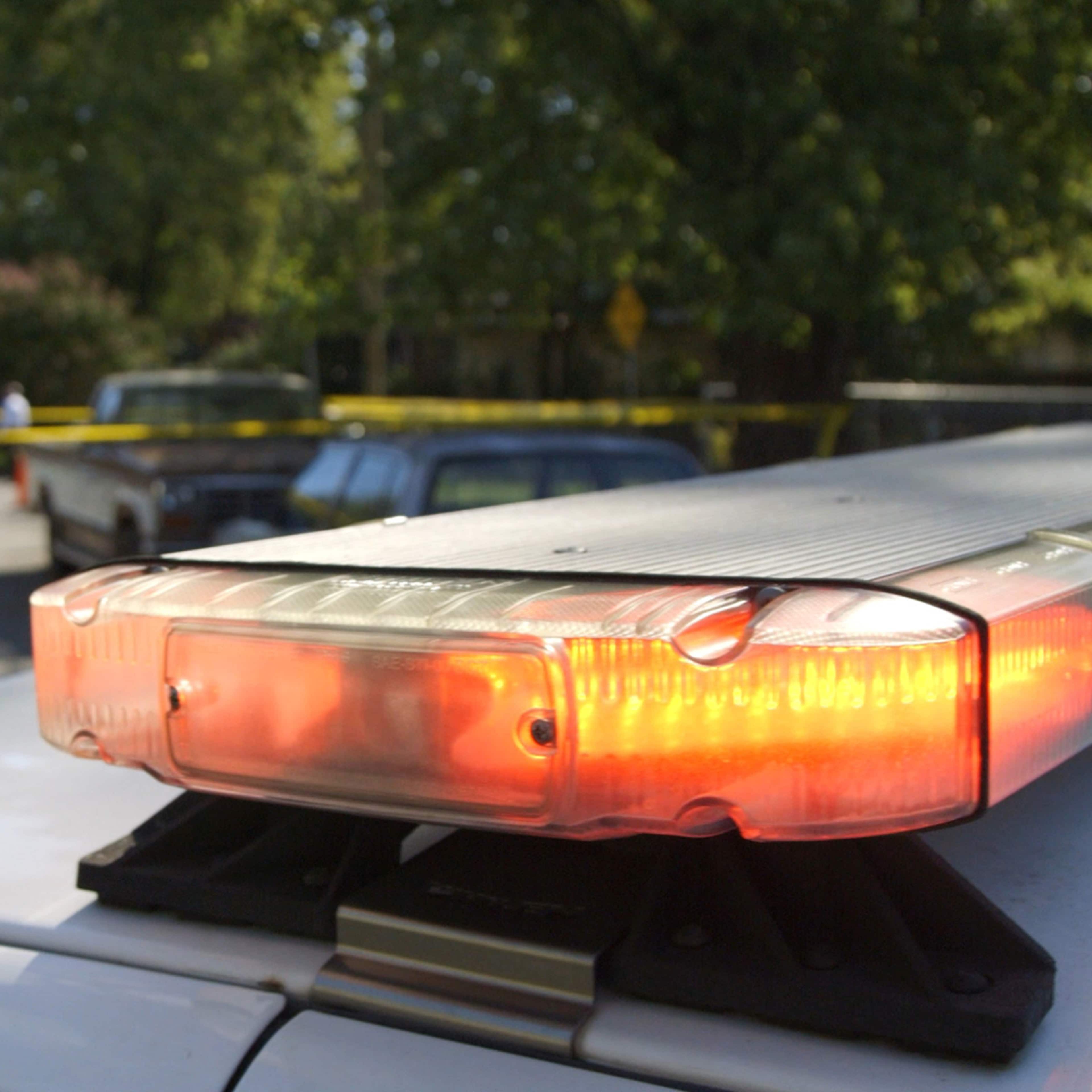 The image shows a police car with its emergency lights flashing, parked on a street surrounded by trees and people in the background.