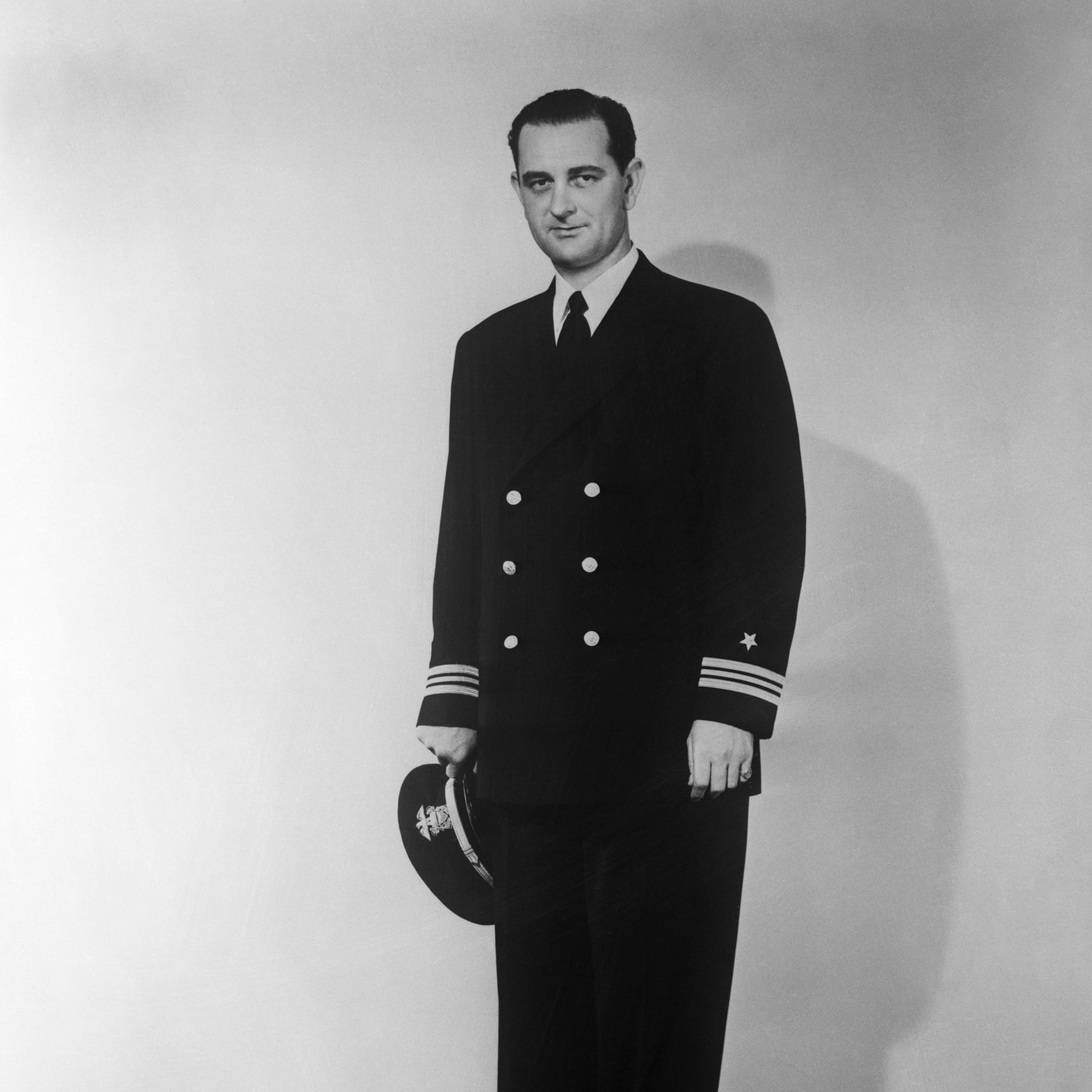 A man in a formal naval uniform stands in a plain studio setting, his gaze directed straight ahead.
