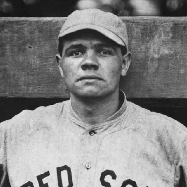 young Babe Ruth, standing against the dugout, wearing a Boston Red Sox uniform
