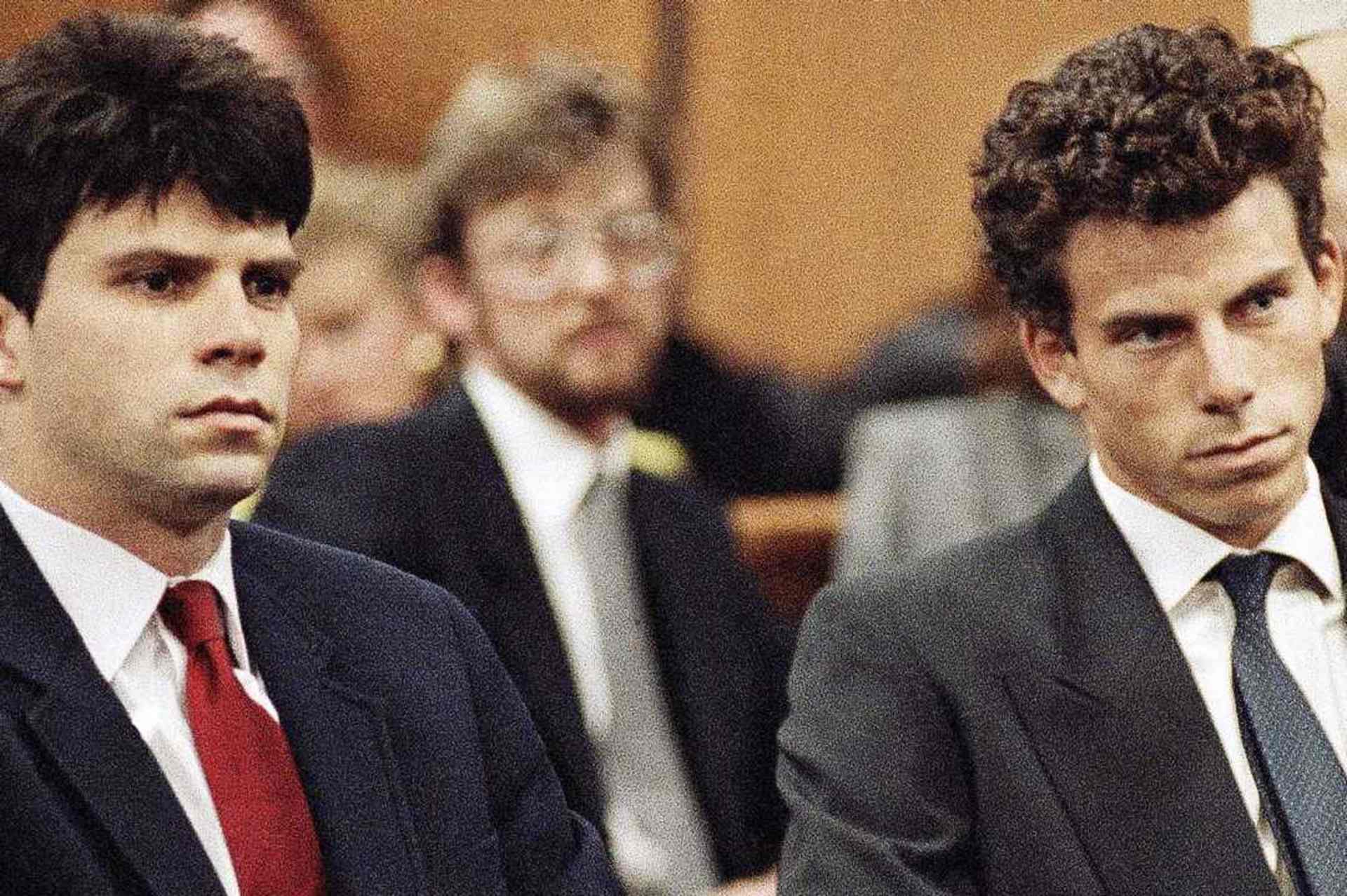 Two young men in suits sitting in a courtroom setting, with other individuals visible in the background.