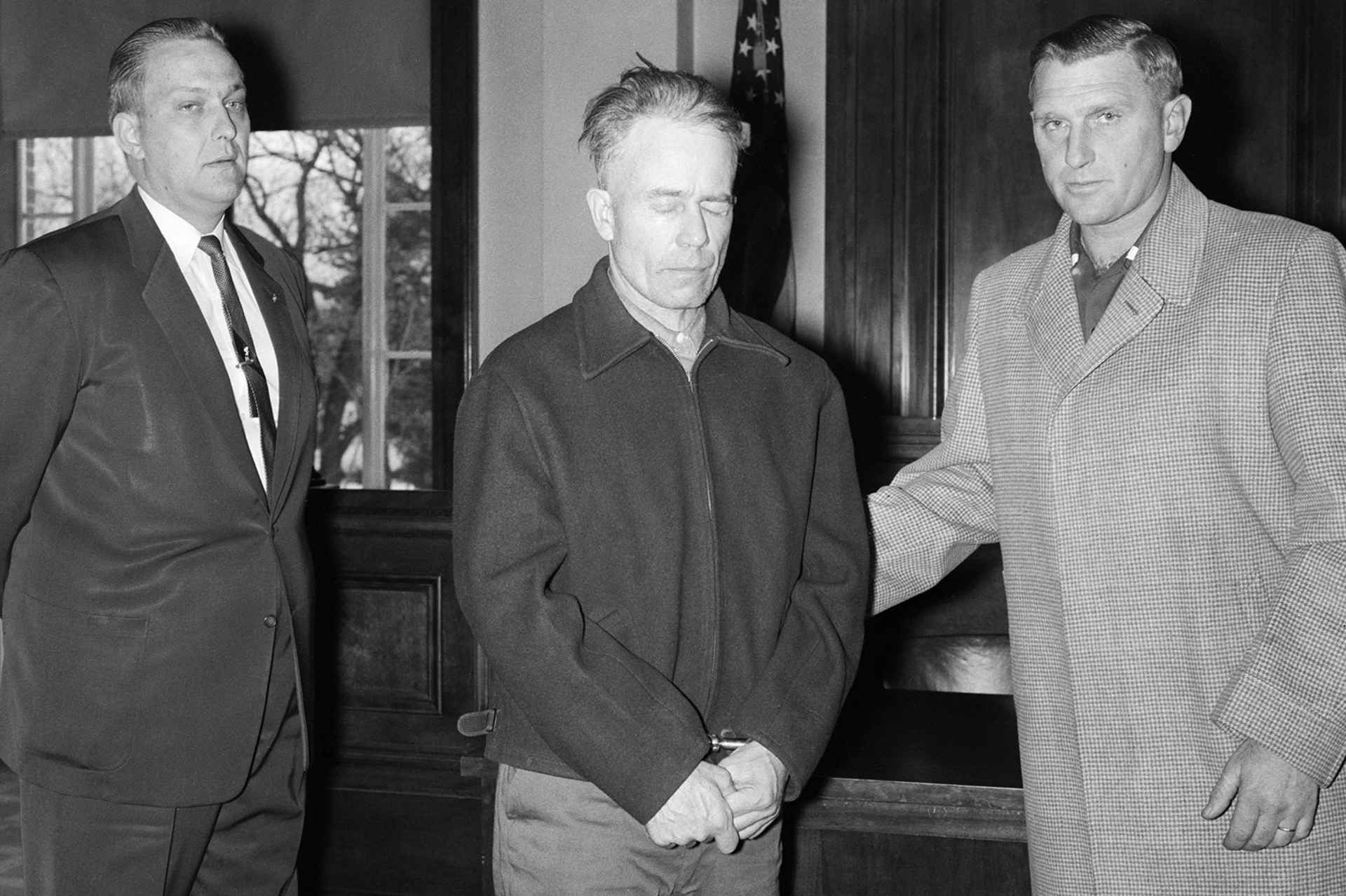 Three men in suits and coats standing in a doorway, with a window visible in the background.