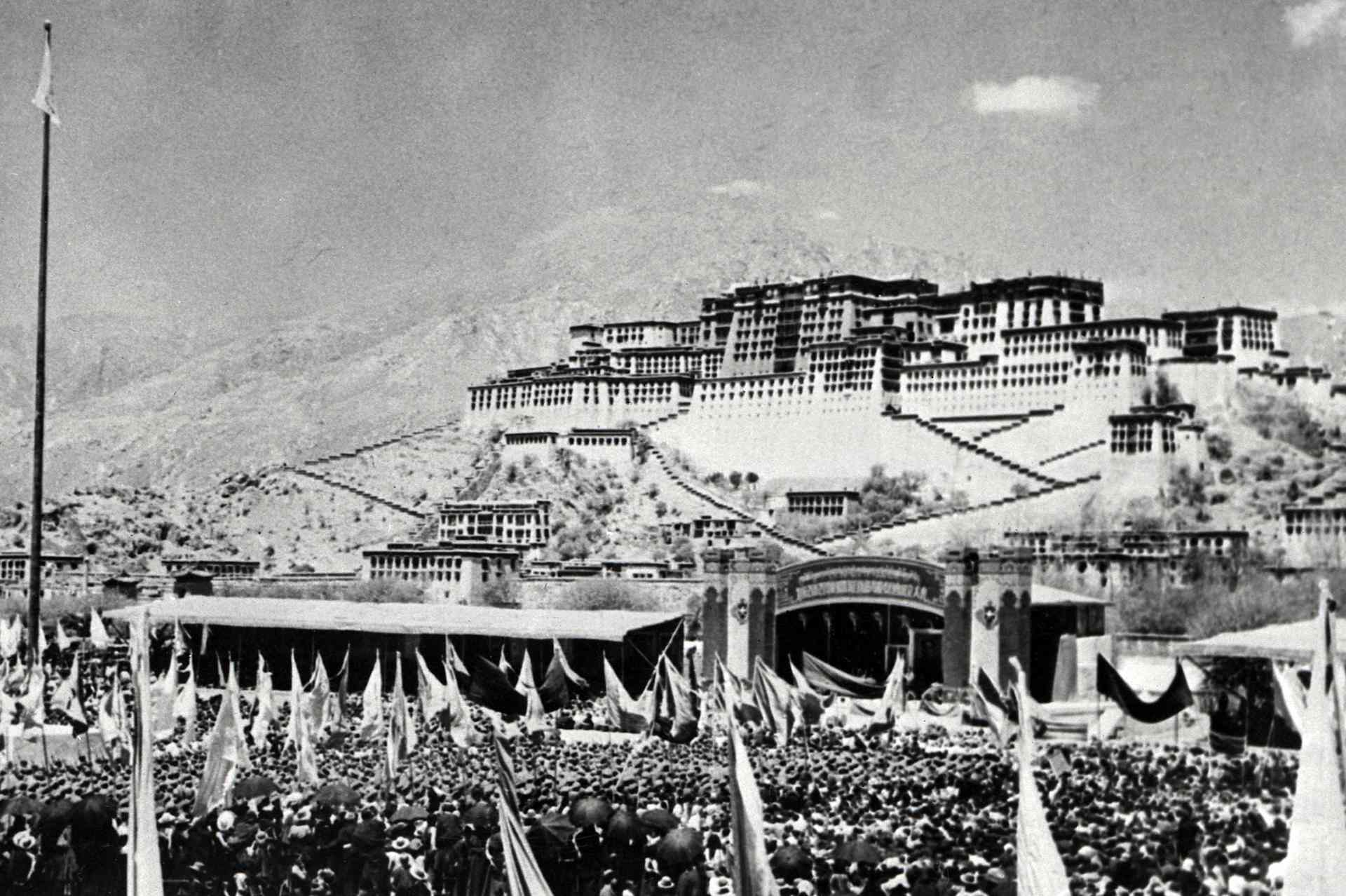 Masses with flags gather beneath a huge Tibetan palace, with mountains behind.