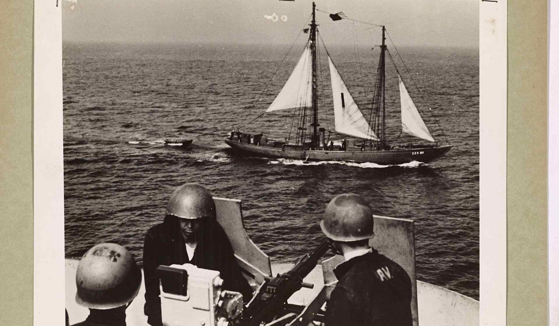 The image shows a Navy anti-aircraft gun crew at battle stations as a Coast Guard auxiliary schooner passes silently by during a patrol. This interaction highlights the close collaboration between Coast Guard and Navy forces during WWII operations.