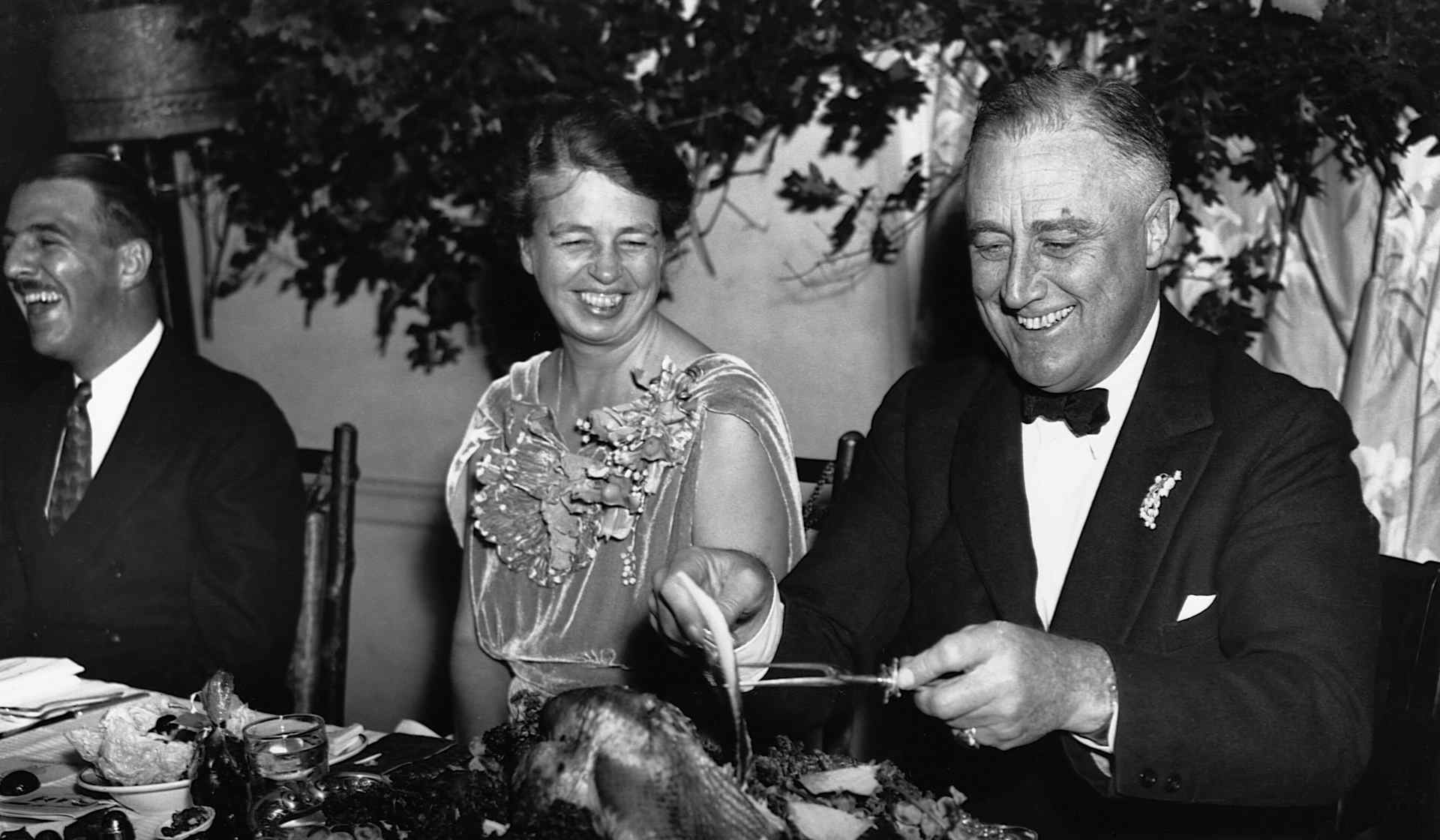 First Lady Eleanor Roosevelt and President Franklin D. Roosevelt smile while sitting at a table as Franklin slices a turkey