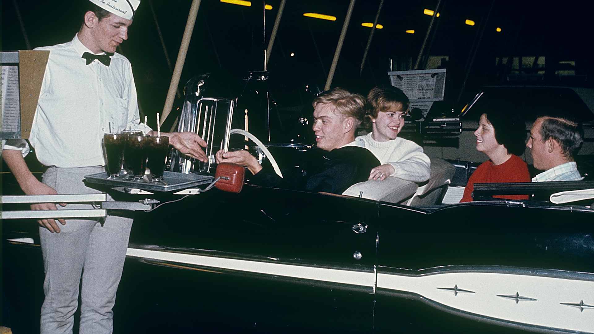 USA in the 1960s. Two young couples in a convertible at a Huddle drive-in restaurant 1964. A waiter is...