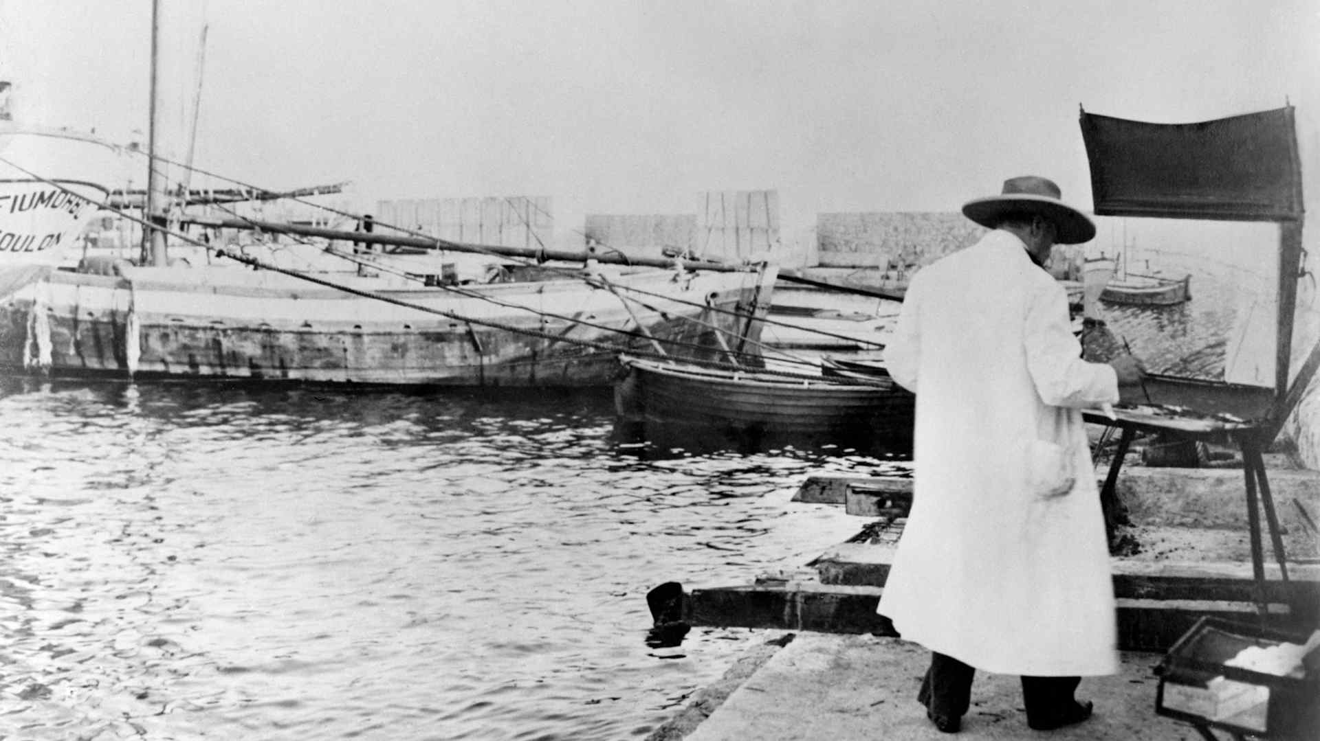 Winston Churchill in a white overcoat and broad-brimmed hat, painting in front of some boats quayside at Toulon, in the south of France, 1946.