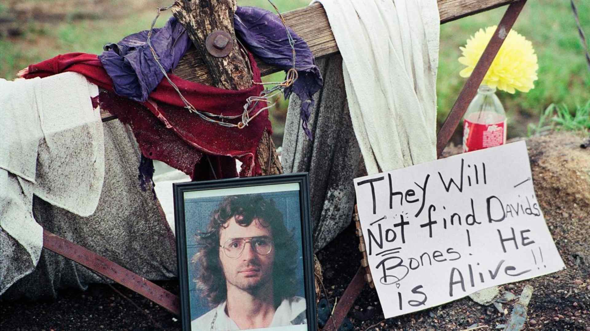 A photo of David Koresh resting beside a wooden cross as part of a monument erected in Waco, Texas by supporters of the Branch Davidian leader and founder. (Credit: NewsBase/AP Photo)