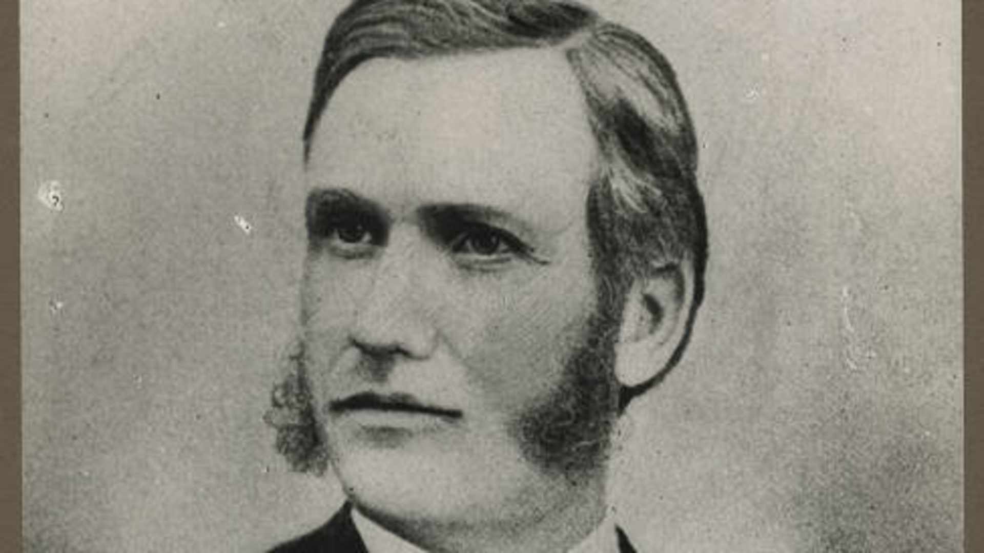 A black and white portrait photograph of a man with a serious expression, wearing a dark suit and tie, set against a plain background.