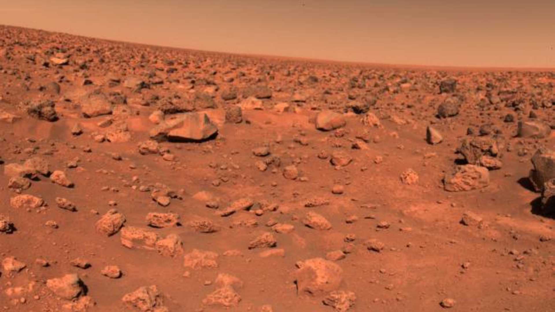 A vast, arid landscape of reddish-brown rocks and soil stretches out under a hazy, orange-tinted sky, creating an otherworldly, Mars-like scene.