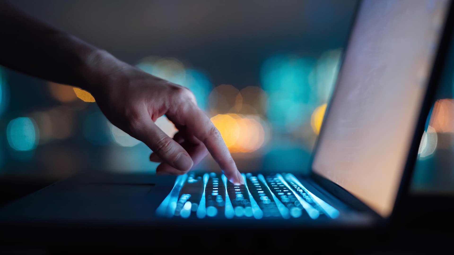 Close up of woman's hand typing on computer keyboard in the dark against colourful bokeh in background, working late on laptop at home