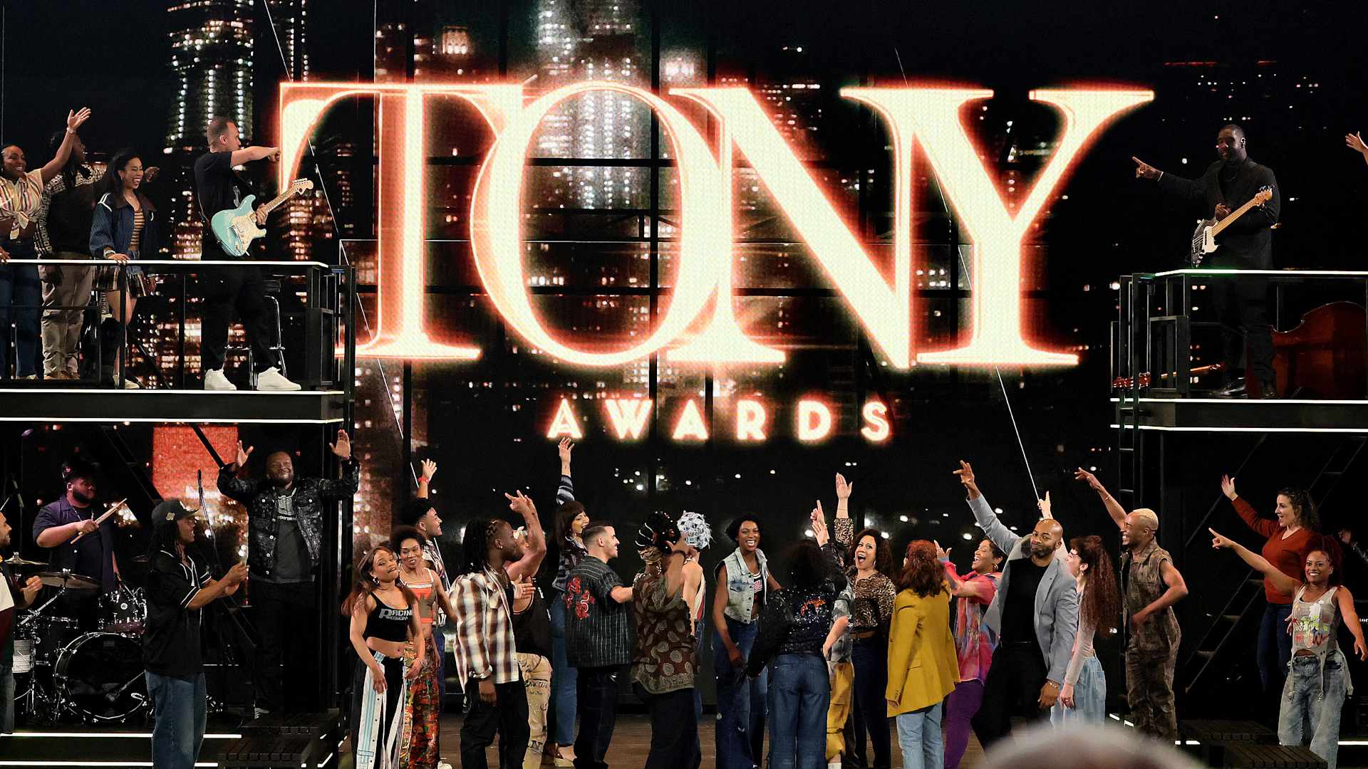 Cast of a Broadway musical on stage pointing upward to the big sign that says 'Tony Awards'