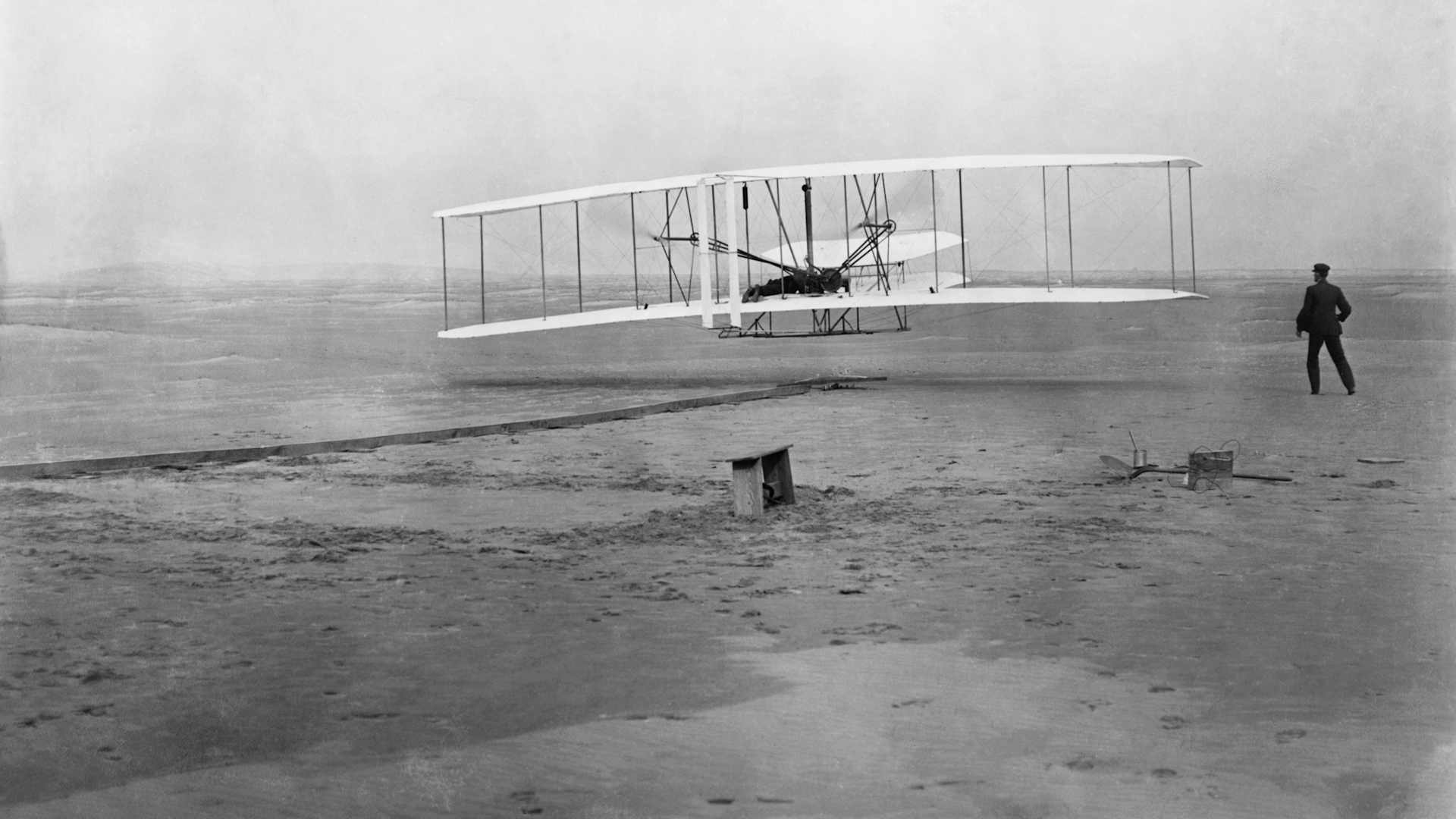 black and white photo of a white biplane on a sandy beach with a man lying in the center and another standing in black to the right of the aircraft.