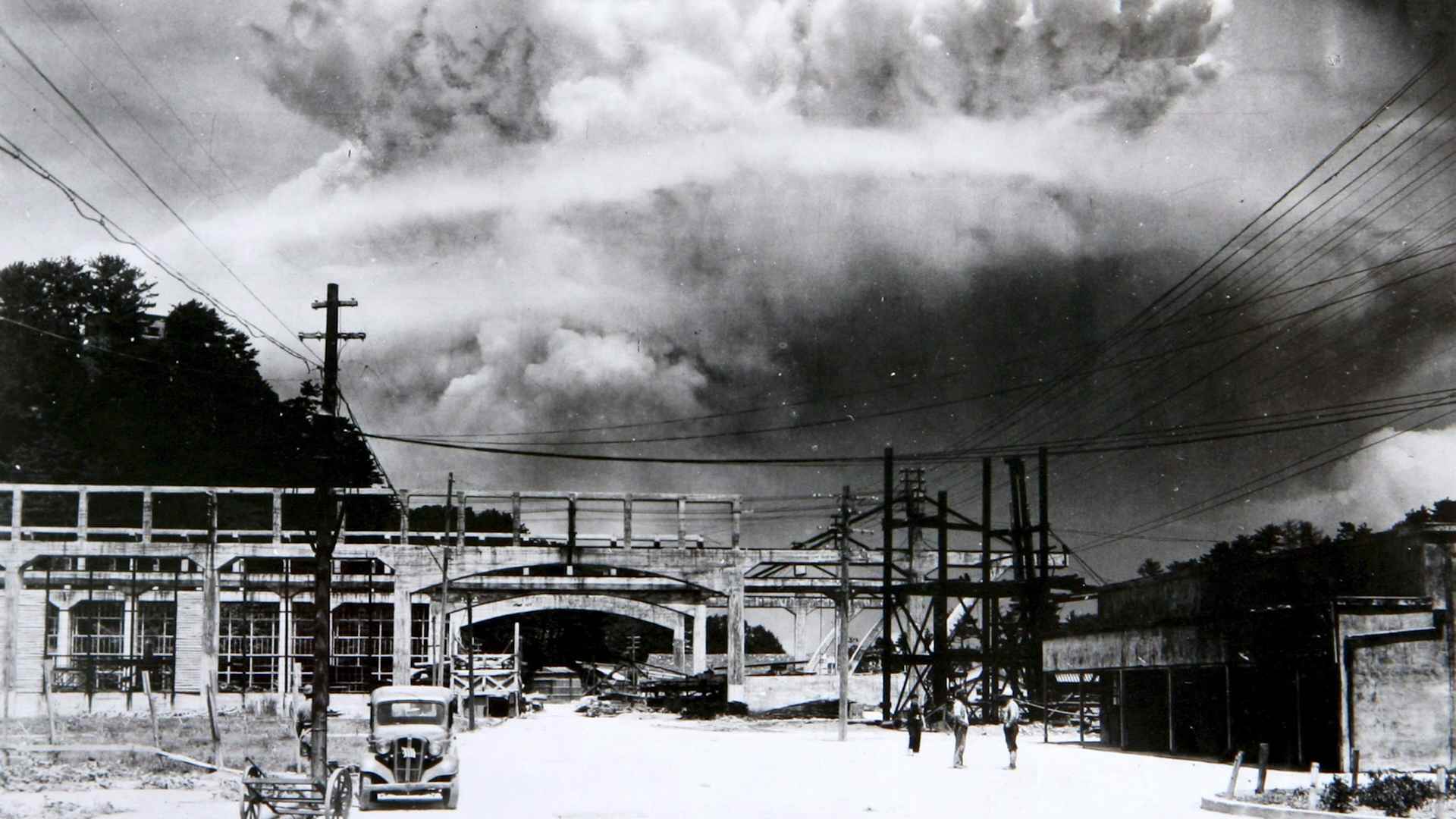 Giant mushroom cloud seen over the city of Nagasaki, with a car and people in the foreground.