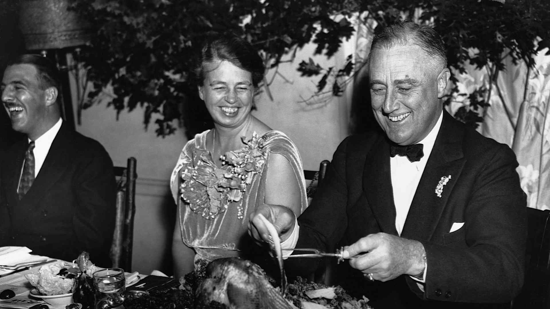 First Lady Eleanor Roosevelt and President Franklin D. Roosevelt smile while sitting at a table as Franklin slices a turkey