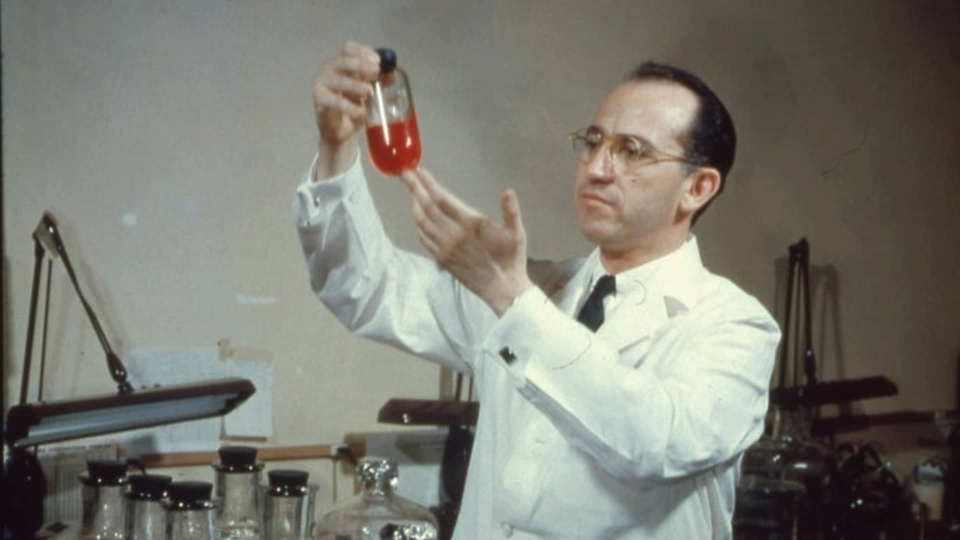 View of American scientist and physician Jonas Salk (1914 - 1995), developer of the polio vaccine, wearing a white lab coat, and smiling while holding up a bottle in the laboratory, mid twentieth century. (Photo by PhotoQuest/Getty Images)