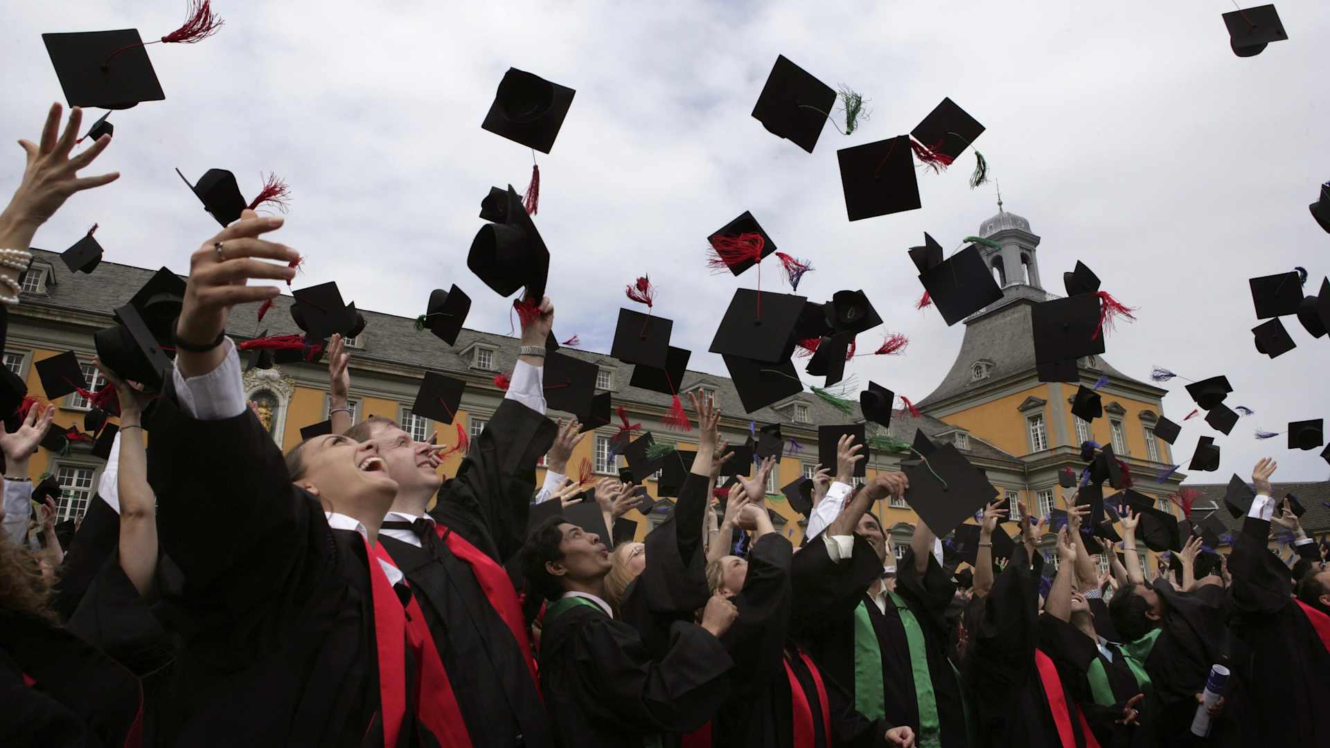 A crowd of smiling graduates in black capes standing outside a university building, throwing their caps in the air.