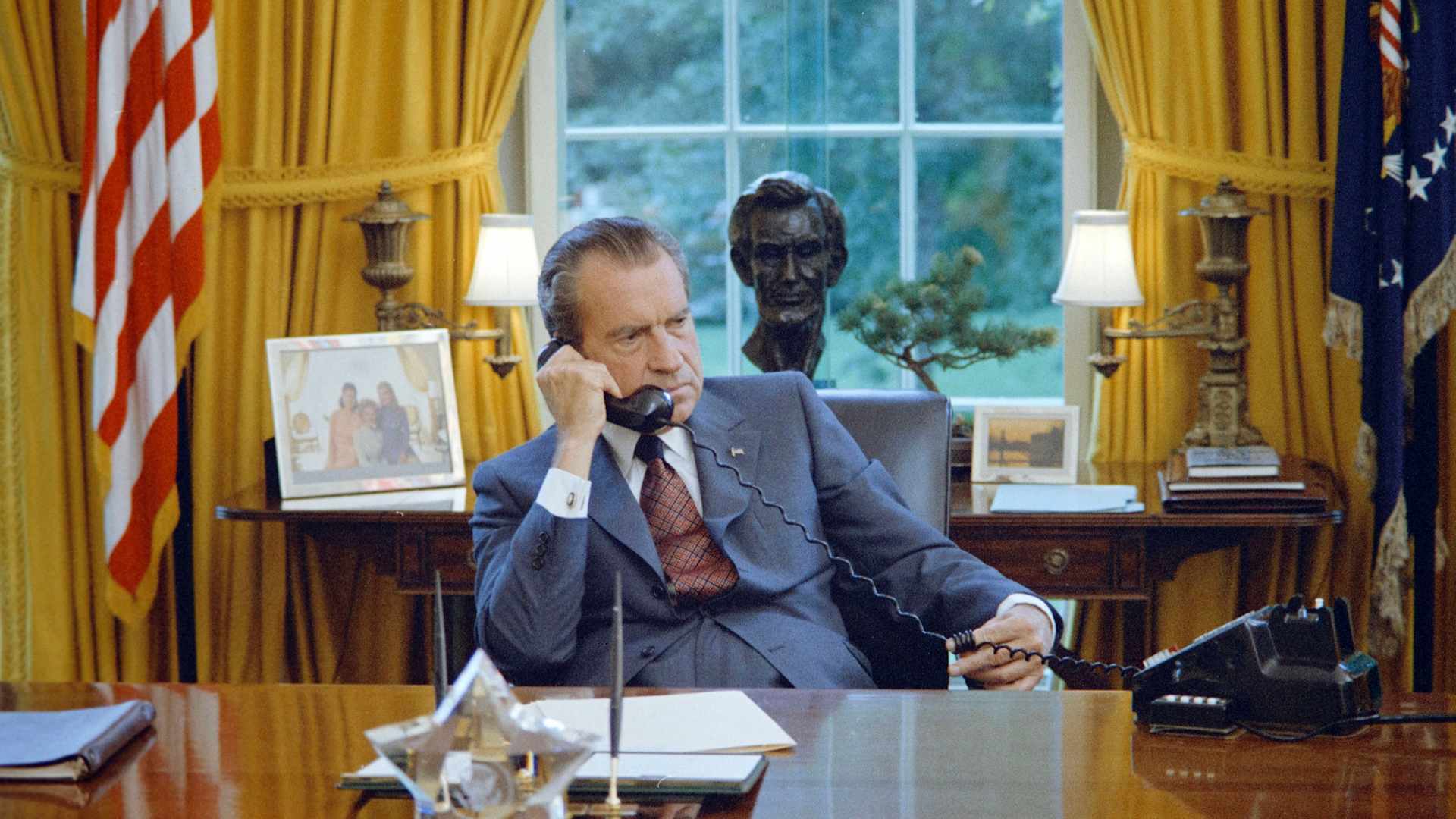 President Richard Nixon in a blue suit, sitting at the desk in the White House Oval Office, talking on the phone, with a bust of Abraham Lincoln behind him in front of a window