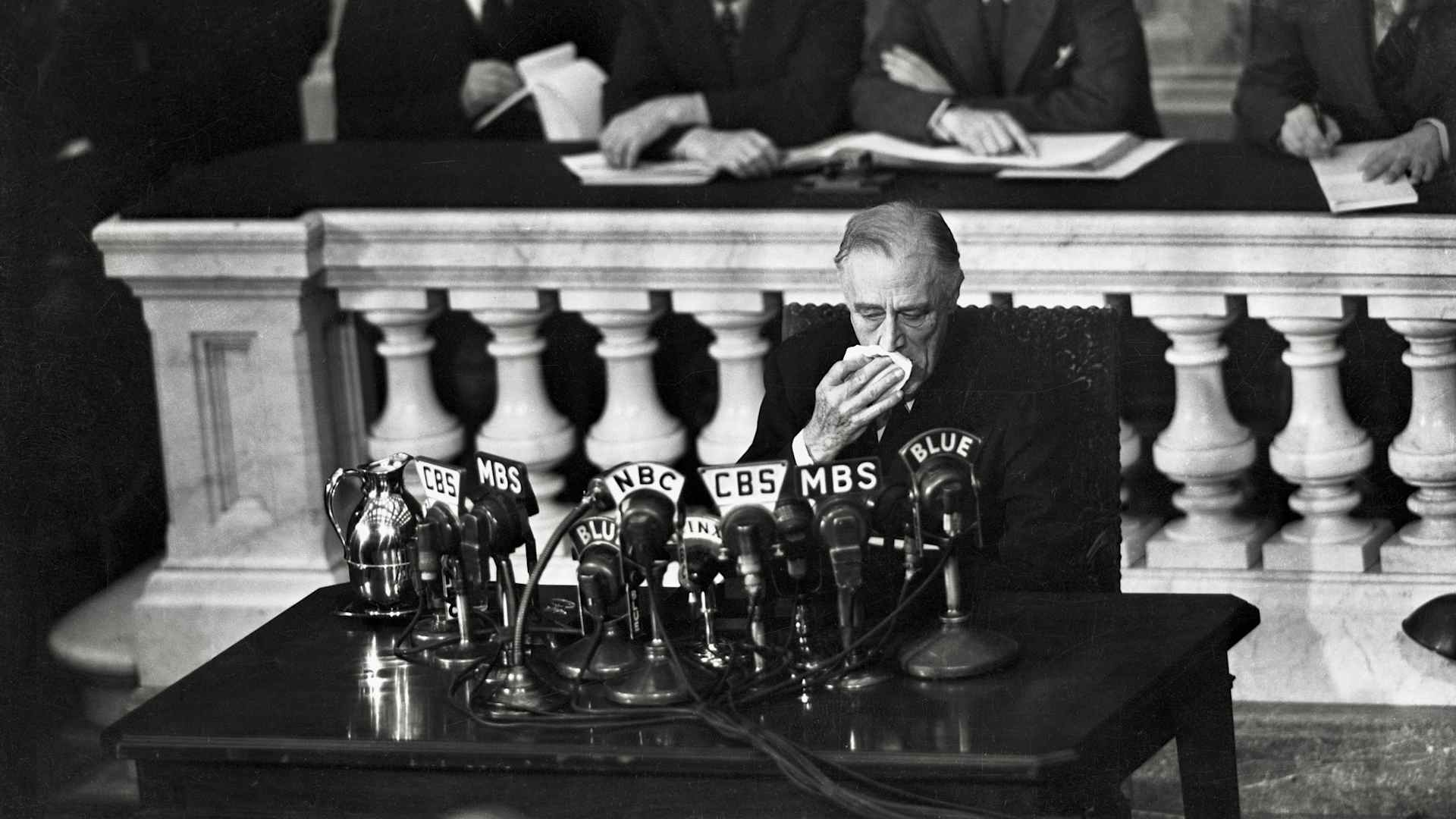 President Franklin D. Roosevelt is shown sitting at a table behind a crowded group of news microphones wiping his mouth with a handkerchief during his reporting to the House of Representatives on the 'Big Three' conference in Yalta.