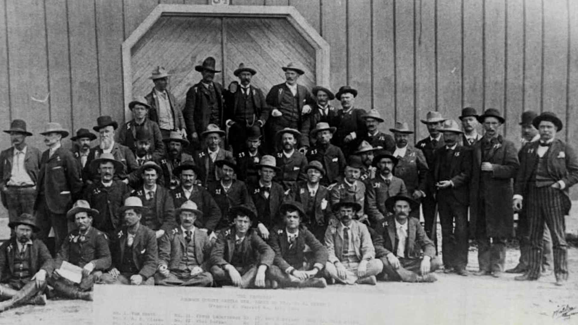 About 40 men in suits and hats posed in front of a barn. After this group of hitmen hired by Wyoming cattle barons to murder settlers were rescued from a siege by the cavalry, they were lined up for this photo. Due to influence from their wealthy backers, the gunmen were all eventually released.